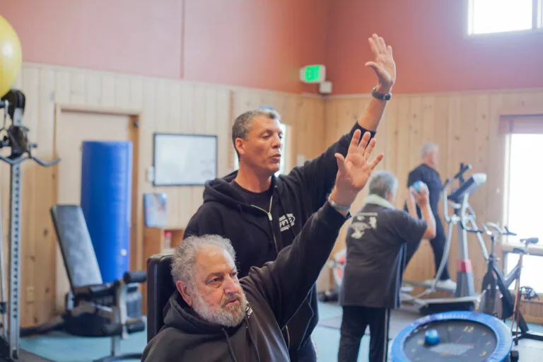 A member stretching one arm overhead while seated in a chair, with a trainer beside him